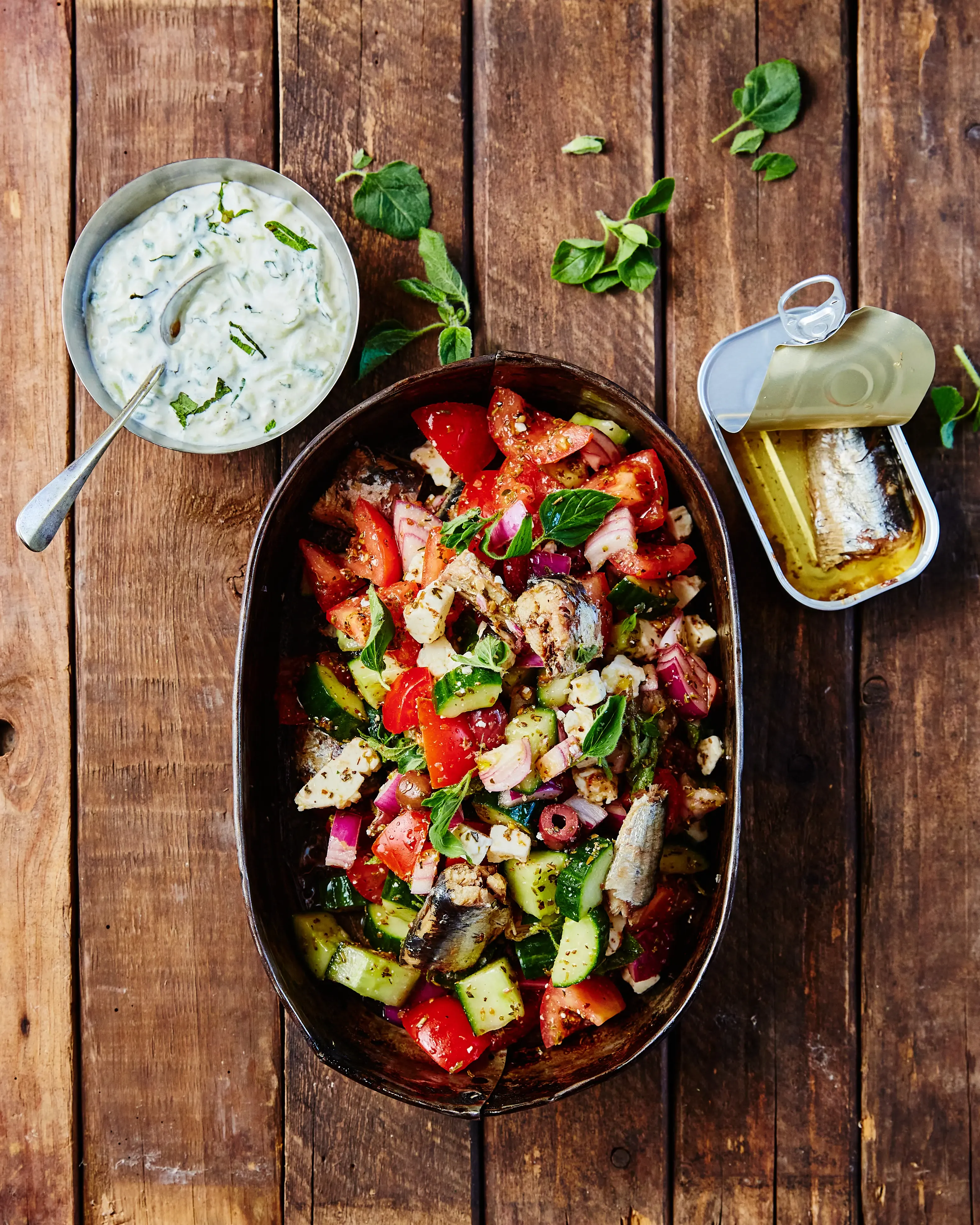 A vibrant Mediterranean salad with octopus, chopped vegetables, creamy dressing, and sardines on a wooden table.