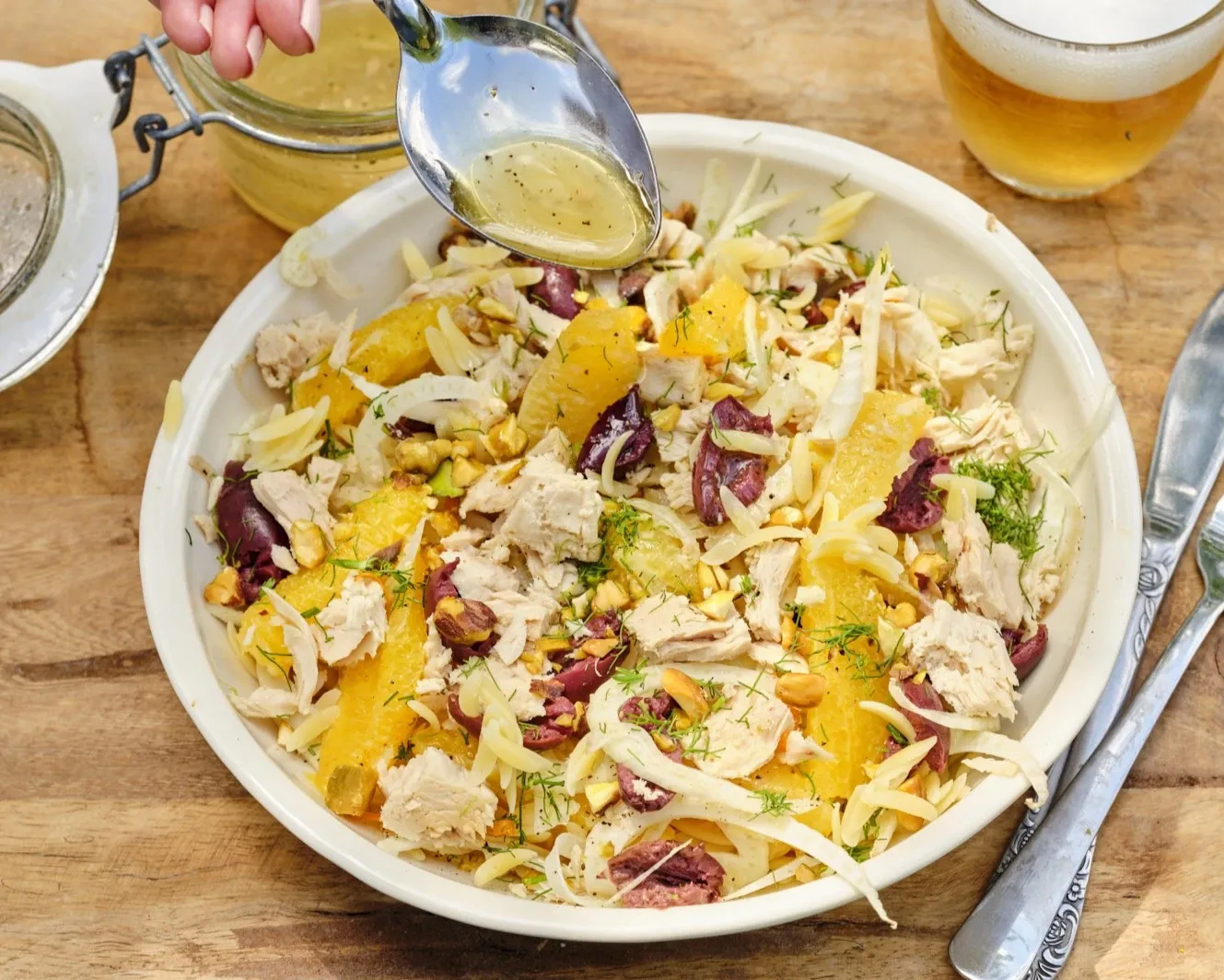 A person pours citrus vinaigrette over a fresh salad with chicken, oranges, olives, and fennel.
