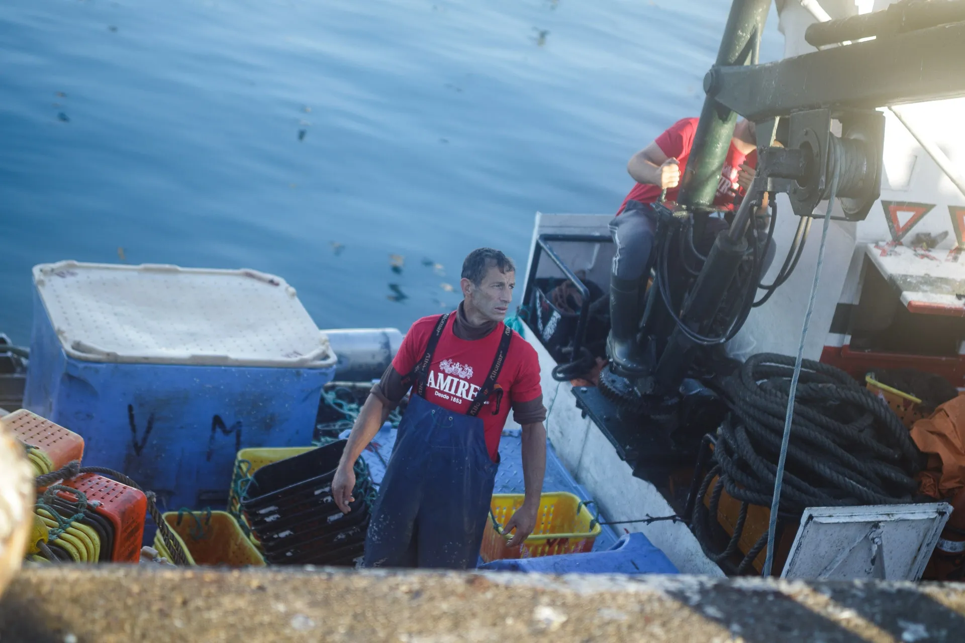 A fisherman in blue overalls and a red shirt stands on a boat with crates.