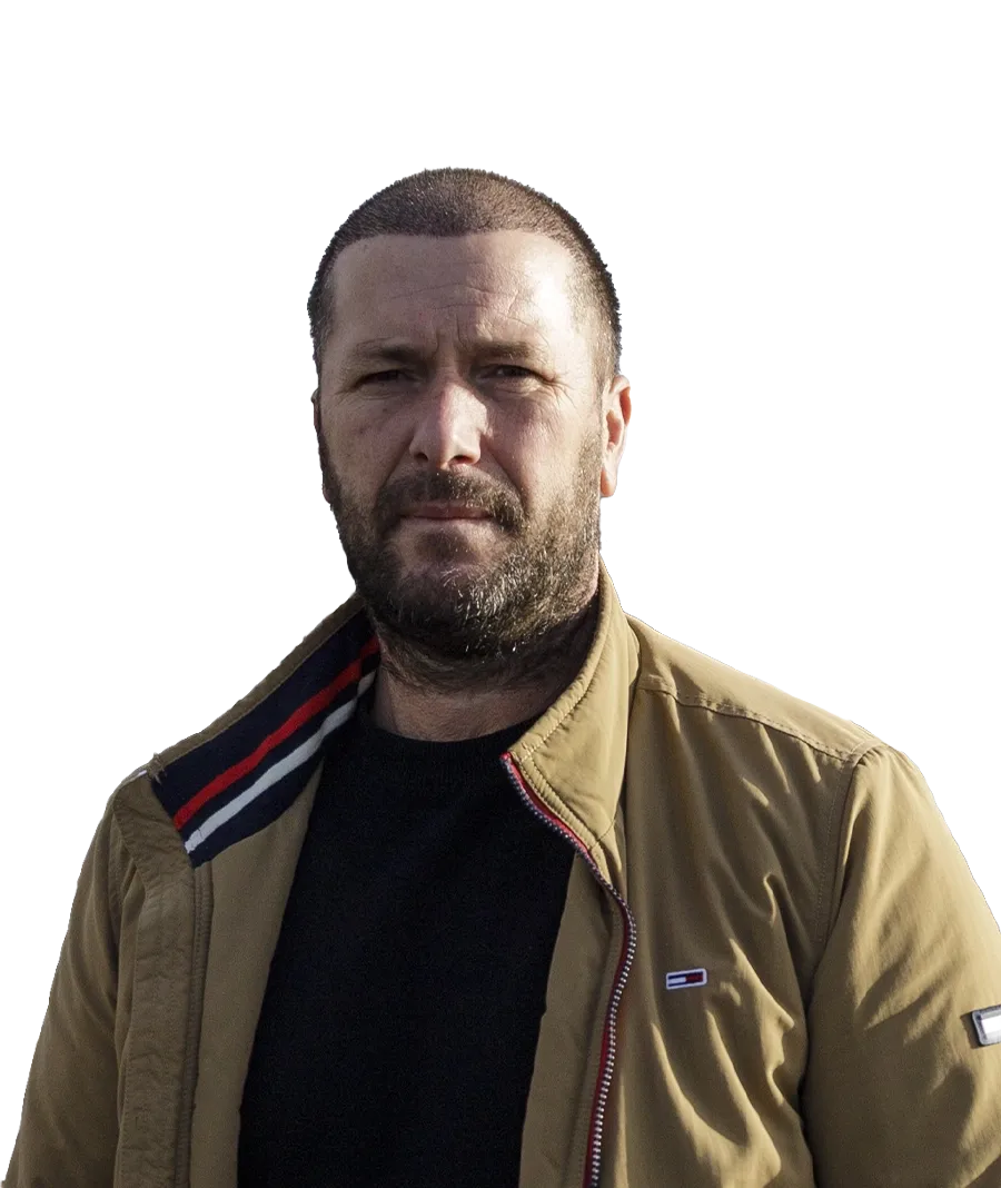 Man with a beard and short hair wearing a tan jacket against a black background.