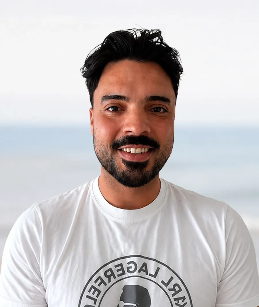A man with dark hair and beard smiles, wearing a white t-shirt outdoors.