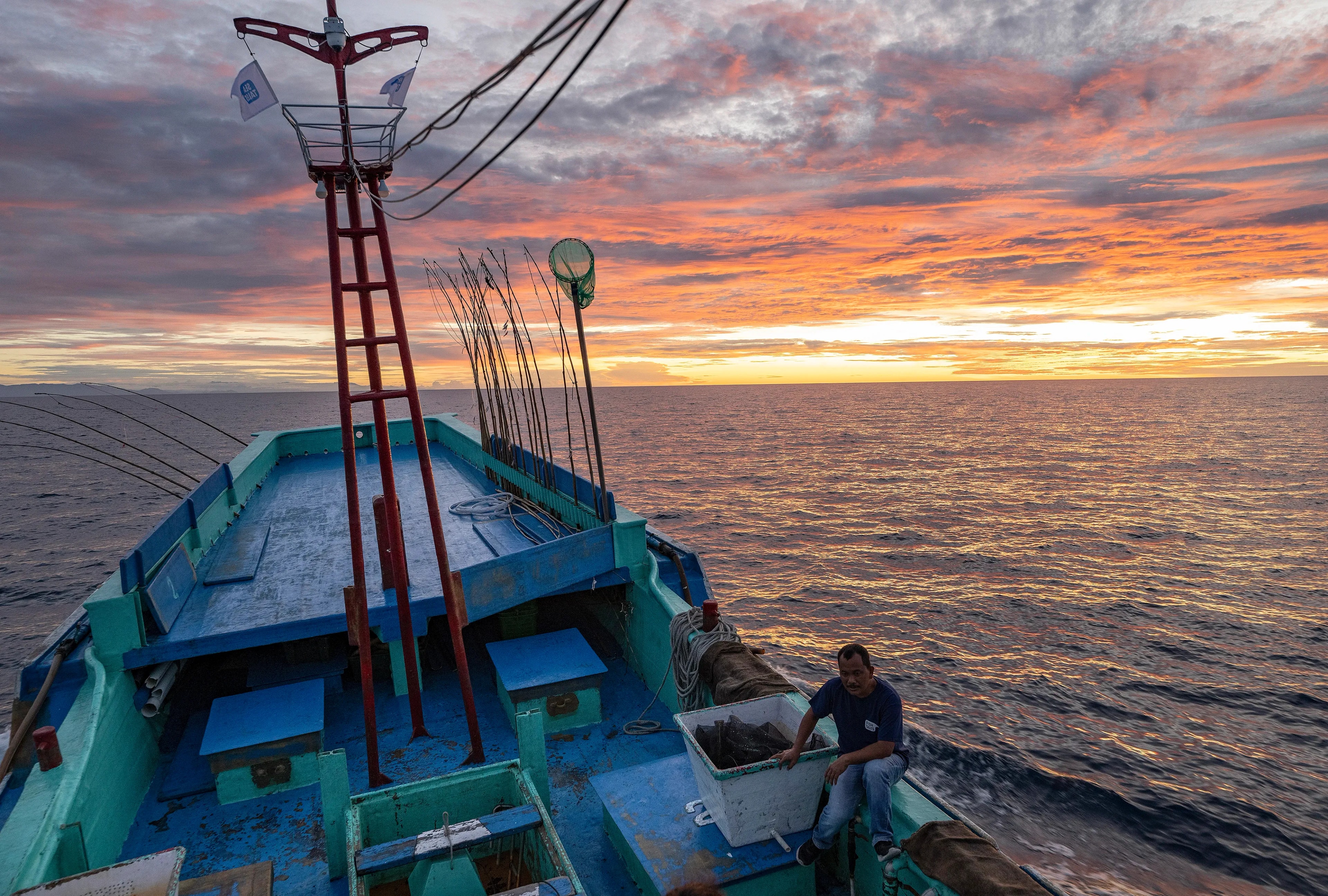 Fishing boat sailing on open ocean at sunrise with colorful sky and a crew member on deck.