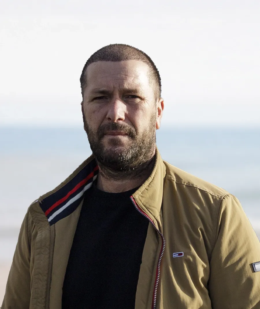 Bearded man in a tan jacket standing outdoors with a blurred ocean background.