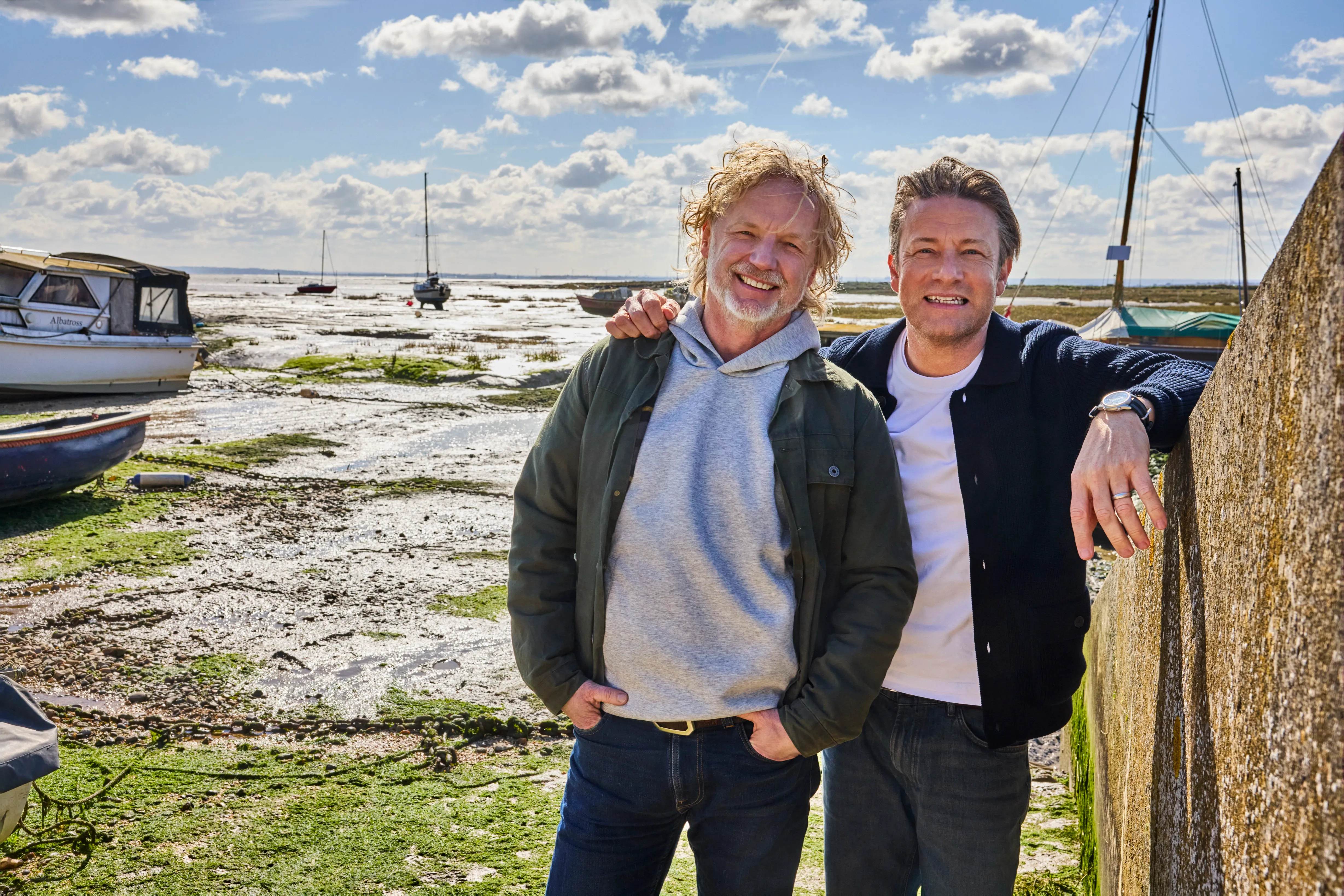 Jamie Oliver and a man stand by a wall overlooking a vast tidal flat with boats.